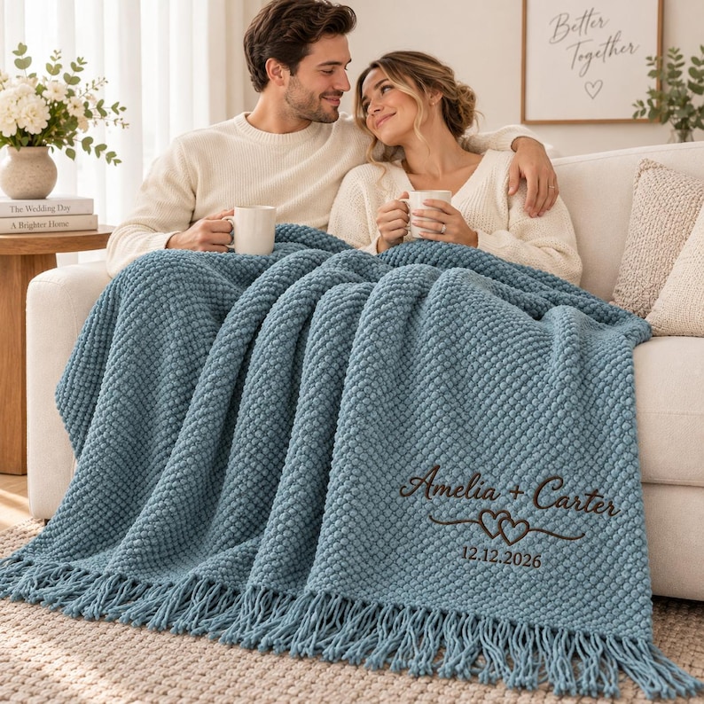May include: A dusty blue textured throw blanket with fringe edges, featuring dark brown embroidered text that reads Amelia + Carter, a double heart graphic, and the date 12.12.2026. The blanket has a raised, bumpy knit pattern throughout its surface.