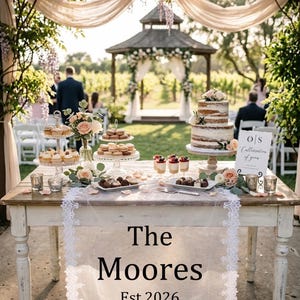 Peut inclure: Une table en bois rustique présente un gâteau de mariage à deux niveaux, des pâtisseries et des desserts. La table est ornée de fleurs et d'un chemin de table en dentelle avec l'inscription "The Moores Est. 2026". La scène se déroule en extérieur, avec un gazebo et des invités en arrière-plan.