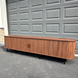 May include: A long, low-profile media console cabinet featuring a rich brown wood grain finish with vertical slat detailing. The unit has rounded ends, two central doors with matching vertical wood handles, and is supported by four thin black metal legs.