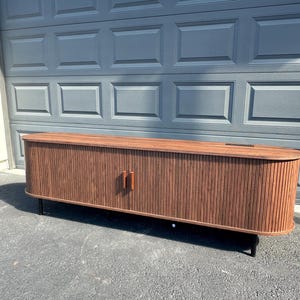 May include: A mid-century modern style wooden cabinet with a dark brown finish. The cabinet has a ribbed texture on the front and curved edges. It features two wooden doors with small, rectangular handles and sits on black metal legs.