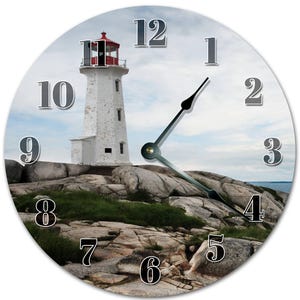 May include: Circular wall clock depicting a white lighthouse with a red top, set against a cloudy blue sky. The clock face has large, grey numerals and black clock hands. The lighthouse is on a rocky landscape with green patches.