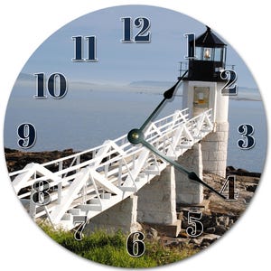 May include: Round wall clock with a coastal scene featuring a white lighthouse and bridge. The clock face has black numerals on a light blue backdrop, with black hands. The lighthouse is set against a seascape, with a bridge leading to it.