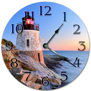 May include: Circular wall clock depicting a lighthouse on a rocky coastline at sunset. The clock face has dark blue numerals and black hands. The lighthouse is white with a red light, set against a blue and orange sky. The clock measures approximately 30 cm in diameter.
