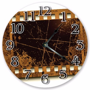 Peut inclure: Horloge murale ronde avec un motif de bande de film. Le cadran de l'horloge présente une bande de film marron et beige avec des chiffres blancs et des aiguilles noires. L'horloge a un fond blanc.