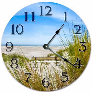 May include: Round wall clock featuring a beach scene with a blue sky, sand, and green grass. The clock face has black hands and large, dark blue numerals. A wooden bench is visible on the beach. The clock is a decorative item.