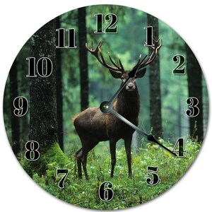May include: Round wall clock featuring a majestic deer in a forest setting. The clock face displays black numerals against a green background, with black clock hands. The deer is brown with large antlers, standing in a lush green forest.