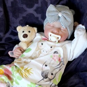 May include: A sleeping baby doll wearing a white and gray striped hat with a bow, a white onesie with a giraffe and zebra print, and a pacifier. The doll is holding a brown teddy bear.