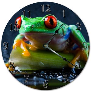 May include: A green tree frog with red eyes sits on a green leaf with water droplets on a black background. The frog is facing the right side of the image. The clock numbers 1 through 12 are visible around the edge of the image.