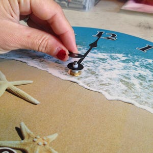 A hand is holding a clock face with a beach scene design, including starfish and waves, against a sandy background.