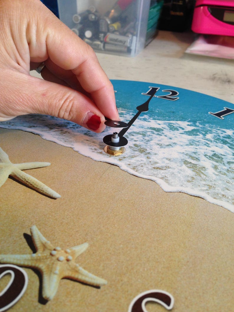 A hand is holding a clock face with a beach scene design, including starfish and waves, against a sandy background.