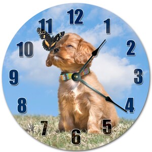 May include: Round wall clock featuring a golden puppy with a butterfly on its nose. The clock face has large, dark blue numbers against a light blue sky background. The puppy wears a colorful collar. The clock hands are black.
