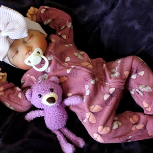 May include: A lifelike baby doll wearing a mauve onesie with a floral pattern, a striped bow headband, and a pacifier. A small purple teddy bear rests beside the doll. The doll is lying on a dark purple surface.
