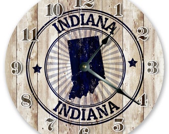Puede incluir: Un reloj redondo de madera con un acabado blanqueado desgastado. La esfera del reloj presenta una silueta azul del estado de Indiana con la palabra "Indiana" escrita en una fuente de estilo vintage. La esfera del reloj también tiene un fondo de veta de madera y marcadores de hora en números romanos.