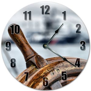 May include: Round wall clock featuring a detailed image of a wooden ship's wheel. The clock face displays black numerals and black clock hands against a blurred background. The wooden wheel has a rich, warm brown color.
