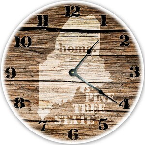 May include: Round wall clock with a rustic wood grain background. The clock face features the outline of the state of Maine, with the words "home" and "Pine Tree State" printed on it. Black numerals and clock hands.