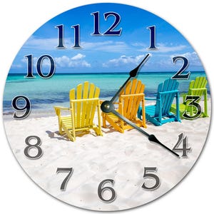 May include: Round wall clock featuring a beach scene with colorful Adirondack chairs on a sandy shore. The clock face displays large black numerals against a backdrop of a blue sky and ocean. The clock hands are black.