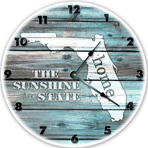 May include: Round wall clock featuring a map of Florida with the word "home" inside the state outline. The clock face has a weathered wood background with the words "The Sunshine State". Black clock hands and numbers.