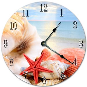 May include: Round wall clock featuring a beach scene with seashells and a red starfish. The clock face displays black numerals and black clock hands. The background shows a beach with blue sky and ocean.