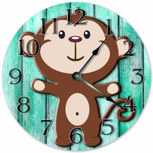 May include: A round wall clock featuring a cartoon monkey design. The monkey is brown with a tan belly and outstretched arms. The clock face has black numerals and hands, set against a teal wooden plank background.