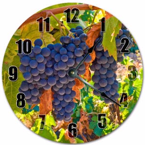 May include: Round wall clock featuring a vibrant image of ripe purple grapes on the vine. The clock face displays black numerals and hands against a backdrop of green leaves and brown vines, creating a naturalistic design.