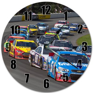 May include: A clock face with a NASCAR race scene as the background. The numbers 1 through 12 are displayed in black on a white background. The race cars are in a tight pack, with the blue car in the lead. The cars are numbered 22, 88, and 5. The clock face is round and has a black border.