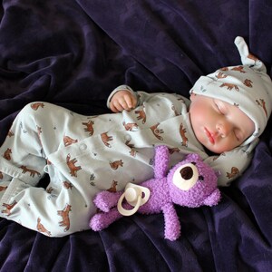 May include: A baby doll wearing a light blue sleeper with a brown animal print and a matching light blue hat is sleeping on a purple blanket. The doll is holding a purple stuffed animal.