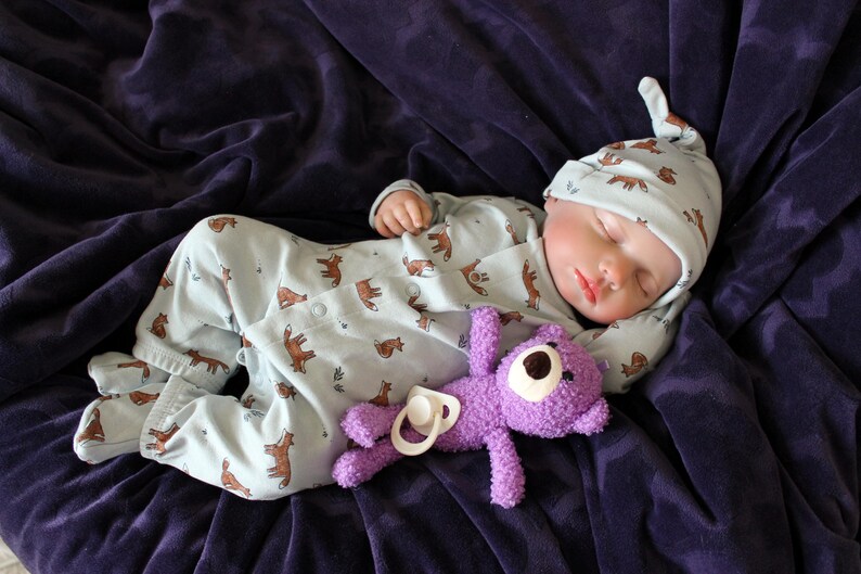 May include: A baby doll wearing a light blue sleeper with a brown animal print and a matching light blue hat is sleeping on a purple blanket. The doll is holding a purple stuffed animal.