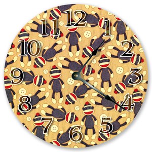 May include: Circular wall clock with a repeating pattern of sock monkey figures. The clock face is tan with white numerals and hands. White buttons are scattered across the design. The sock monkeys are dark grey with red and white accents.
