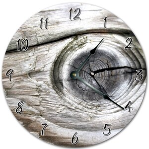 Peut inclure: Horloge murale ronde au design rustique. Le cadran de l'horloge présente un gros plan de bois vieilli avec un nœud au centre. Des chiffres noirs indiquent les heures et des aiguilles noires indiquent l'heure.