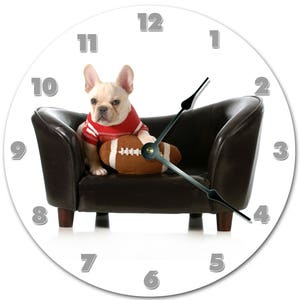 May include: Round wall clock featuring a light-colored French Bulldog wearing a red and white striped shirt, seated on a small brown leather couch. The dog is holding a brown football. The clock has silver numerals and hands.