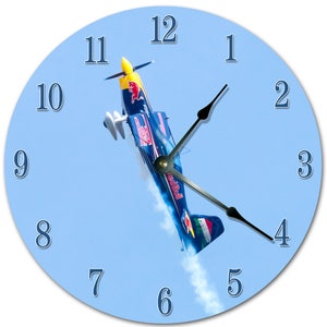 May include: A blue and red airplane with the word "Red Bull" on the side is flying upside down against a light blue sky. The airplane is part of a clock with black hands and white numbers.
