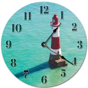 May include: Round wall clock featuring a lighthouse in the ocean. The lighthouse is red and white, with a red top. The clock face is a light blue with black numbers and clock hands.