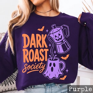 May include: A dark purple long-sleeve sweatshirt featuring a graphic print with the text "DARK ROAST society" in orange. The design includes a light purple moka pot with a jack-o'-lantern face pouring liquid into a ghost-shaped mug with bats.