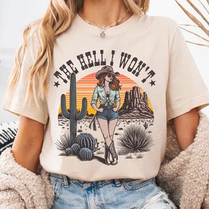 Peut inclure: T-shirt beige avec un motif de femme en chapeau et bottes de cowboy, et le texte "THE HELL I WON'T." Le design comprend un paysage désertique avec des cactus et des montagnes, dans des tons orange, marron et vert.