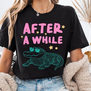 May include: Black t-shirt with the text "AFTER * A WHILE" in pink, with yellow star accents. Below the text is a teal alligator wearing sunglasses. The shirt is a casual style.