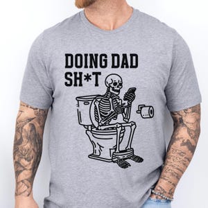 Può includere: T-shirt grigio chiaro con la scritta "DOING DAD SH*T" sopra un'illustrazione nera di uno scheletro seduto su una toilette che guarda un telefono. Un rotolo di carta igienica è a destra dello scheletro.