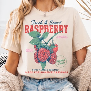 May include: Beige t-shirt with a graphic of red raspberries, green leaves, and the text "Fresh & Sweet RASPBERRY." Additional text includes "Locally Grown," "Picked at Peak Season," "Always Fresh," and "Sweet Little Berries Made for Summer Cravings."