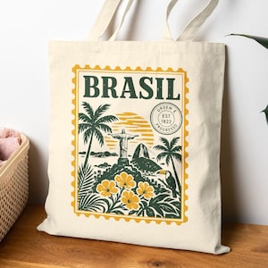 May include: A cream-coloured canvas tote bag featuring a rectangular stamp-style graphic. The print displays the word BRASIL, the Christ the Redeemer statue, Sugarloaf Mountain, palm trees, yellow flowers, a toucan, and a circular seal.