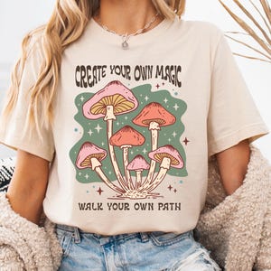 以下が含まれることがあります： ベージュのTシャツ。カラフルなキノコのグラフィックが特徴で、「CREATE YOUR OWN MAGIC」と「WALK YOUR OWN PATH」の文字がデザインされています。ピンクとオレンジのキノコが緑色の背景に描かれています。