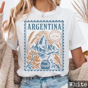Op de afbeelding: Een wit t-shirt met een blauw en beige grafisch ontwerp in de vorm van een postzegel. De print toont het woord ARGENTINA, een bergketen, een vogel, een mate-beker met een rietje en de tekst EN UNION Y LIBERTAD EST 1816.