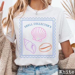 May include: A white short-sleeve t-shirt featuring a blue rectangular stamp-style border. Inside the border, the text "SHELL COLLECTOR'S" appears in blue above three line-art illustrations of seashells in pink, purple, and orange colors.