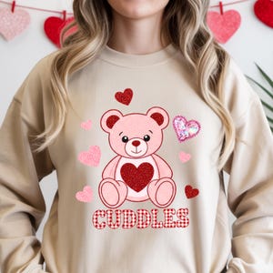 Könnte beinhalten: Beiges Sweatshirt mit einem rosa Teddybären-Design, das ein rotes Herz und rosa Herzen zeigt. Das Wort "CUDDLES" ist in einem rot-weiß karierten Muster aufgedruckt. Dekorative Herzen sind im Hintergrund zu sehen.
