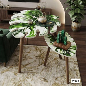 May include: A set of two nesting tables with white and green leaf-shaped tops and brown wooden legs. The larger table holds a metallic teapot and a small bowl, while the smaller table features three green crystal points on a rectangular book.