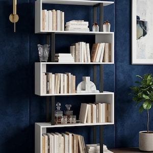 May include: A modern white bookshelf with black metal supports, filled with books and decorative items. The shelves are staggered and create an interesting visual design. The background is a dark blue wall.