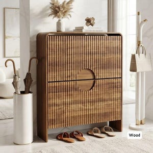 May include: A rectangular wooden shoe cabinet with a vertical fluted texture and a central circular cutout handle. The cabinet stands on a floor near a white cylindrical umbrella stand containing two umbrellas, a white handbag, and two pairs of sandals.