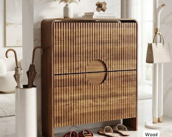 May include: A rectangular wooden shoe cabinet with a vertical fluted texture and a central circular cutout handle. The cabinet stands on a floor near a white cylindrical umbrella stand containing two umbrellas, a white handbag, and two pairs of sandals.
