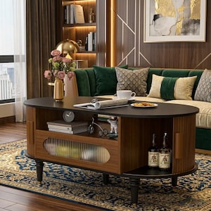 May include: An oval-shaped coffee table with a dark top and a lower shelf. The table has a wooden frame with a ribbed glass door and two bottles on the bottom shelf. The table is in a living room with a sofa and rug.