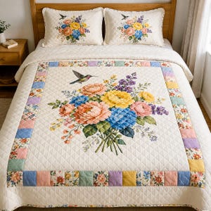 Pode incluir: Conjunto de edredão floral creme com um grande buquê de flores coloridas, incluindo flores rosa, amarelas e azuis. O edredão tem um padrão de diamante e uma borda com patchwork floral. Duas fronhas combinando com desenhos florais e beija-flores completam o conjunto.