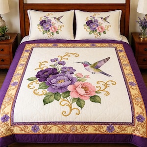 May include: A quilted bedspread and two pillow shams featuring a floral and hummingbird design. The bedspread has a white center with a large floral arrangement in shades of purple and pink. The border is purple with a gold floral pattern.