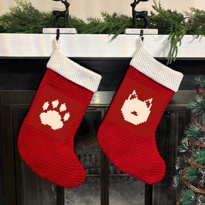 May include: Two red Christmas stockings with white cuffs hang from a mantel. One stocking features a white paw print, the other a white pixelated dog face. Greenery and deer figurines decorate the mantel.